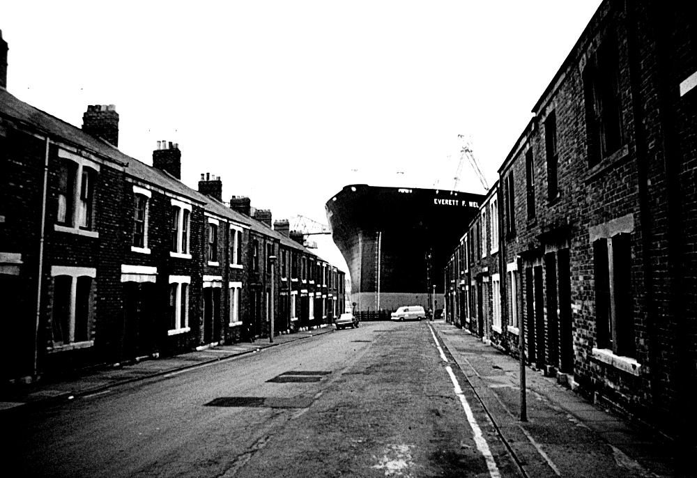Black and white photograph of large ship seen at the end of a street of terraced houses